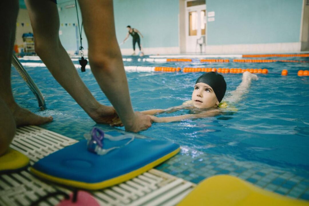 Criança aprendendo a nadar em piscina com ajuda de uma instrutora.